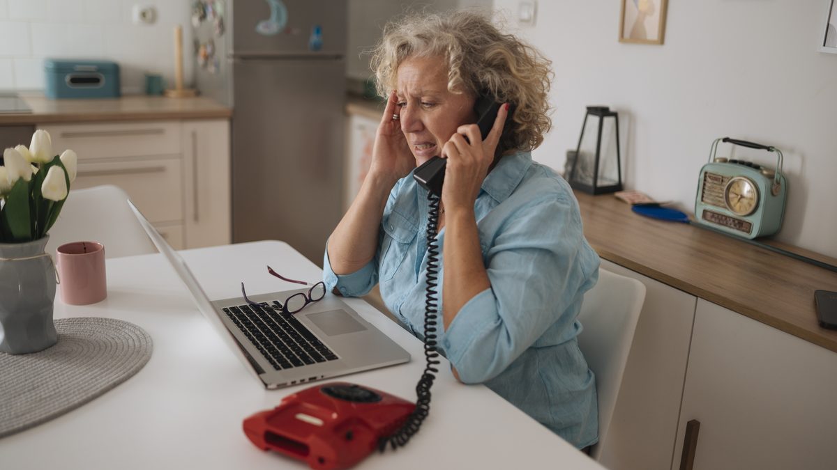 An older woman looking concerned while talking on a landline phone at her kitchen table with a laptop open